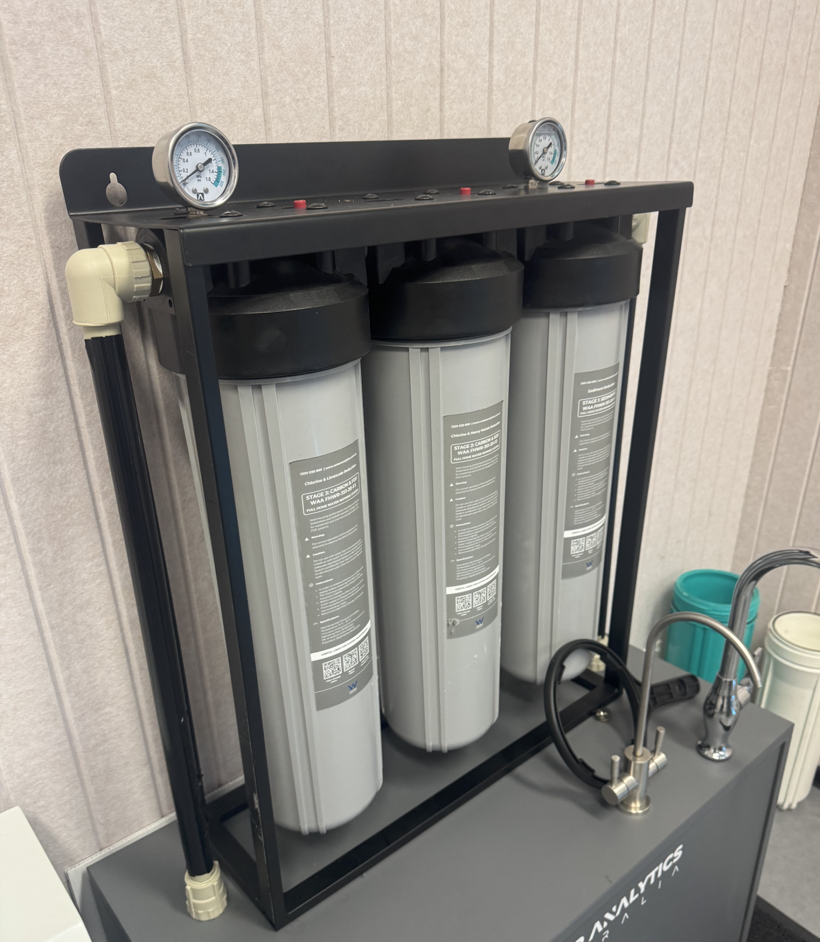 Three-stage whole-home water filtration system with pressure gauges and a chrome mixer tap mounted on display stand